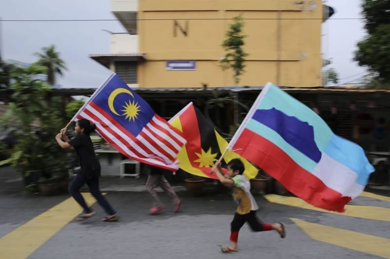 Three children march along a street, each with a flag: Malaysia (left), Sarawak (centre), Sabah (right), against a multi-storey building.